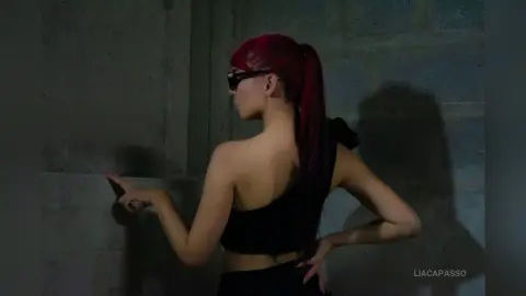 Woman with red hair in a black strapless top posing against a concrete wall in moody lighting.
