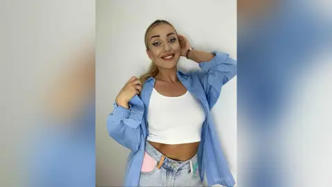 Smiling blonde woman in a white crop top and blue button-down shirt posing against a white background.