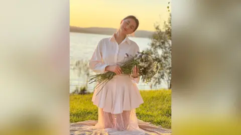 Dreamy woman in white linen outfit holding a wildflower bouquet by a lake at sunset.