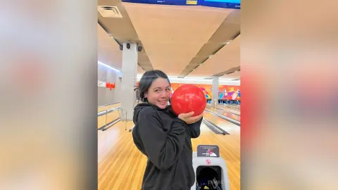Smiling woman in a black hoodie holding a red bowling ball at a bright indoor bowling alley.