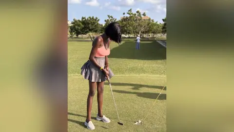 Young woman in a pink tank top and grey skirt practicing golf on a sunny green course.