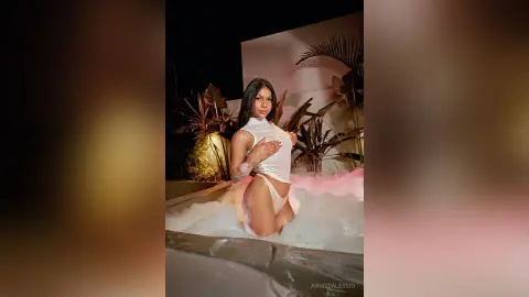 Glamorous woman in a white bodysuit relaxing in a bubbly outdoor jacuzzi at night.