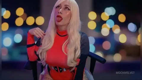 Blonde woman in a red Deadpool cosplay outfit sitting against a blurred city bokeh background at night.