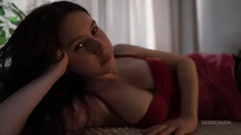 Young woman in a red dress relaxing on a bed in a soft, moody indoor setting.