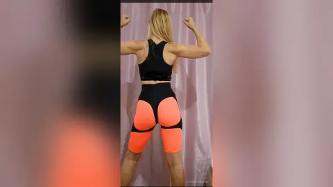MonikaSwenson — Fit blonde woman in black sports bra and orange gym shorts flexing muscles in front of a white curtain.