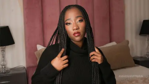 AmaraHoffman poses in a black turtleneck sweater with her long braids draped over her shoulders in a bedroom setting.