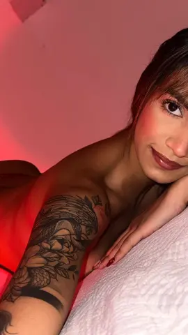Smiling woman with floral arm tattoos relaxing on a white bed in warm red lighting.