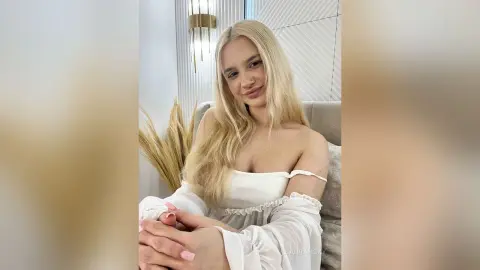 Blonde woman in a white off-the-shoulder blouse smiling in a bright, minimalist indoor setting.