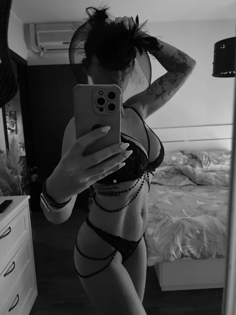 MiaFit poses with a direct gaze, wearing large glasses and showing her dark hair and bare shoulders against a plain brown wall.