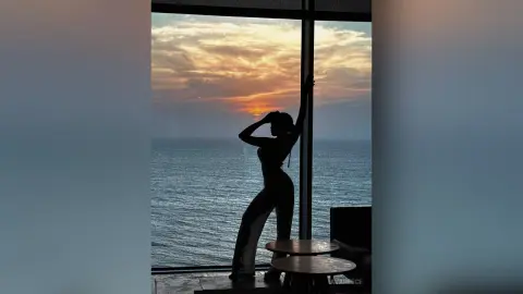 Silhouette of a woman in white trousers posing by a window overlooking a sunset ocean view.
