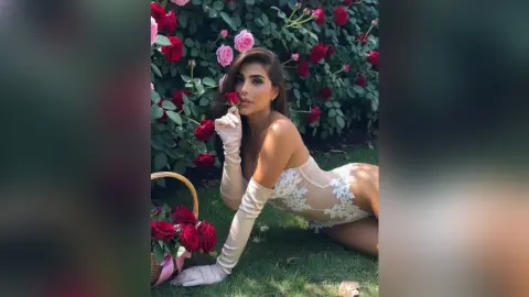 AllishaCoral — Elegant woman in white lace bodysuit and long gloves posing in a lush rose garden.
