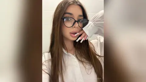 Young woman with long brown hair and glasses wearing a white button-down shirt in a bright indoor setting.