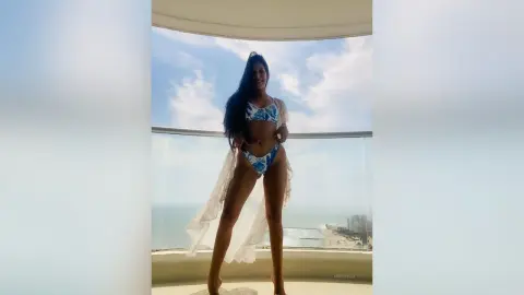 Woman in blue floral bikini and sheer white cover-up standing on a sunny balcony overlooking the ocean.