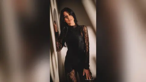 NerissaMist poses in a black lace bodysuit with long sleeves, leaning against a wall in soft lighting.