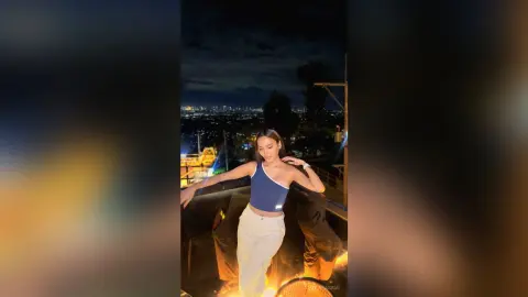 Young woman in a blue bodysuit and white trousers posing on a rooftop at night with a city skyline view.