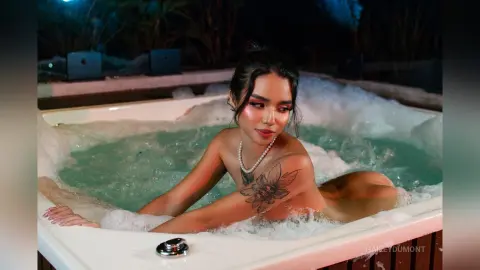 Young woman with tattoos relaxing in a bubbling outdoor hot tub at night under moody lighting.