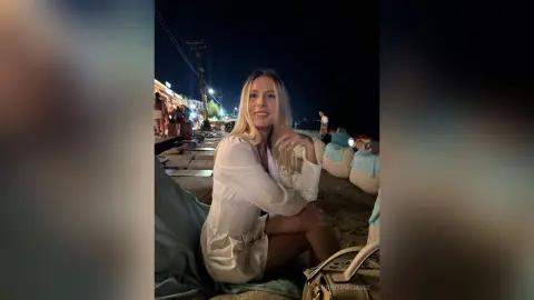 ChristineDavis smiles while sitting outdoors at night, wearing a white long-sleeved shirt and showing her bare legs.