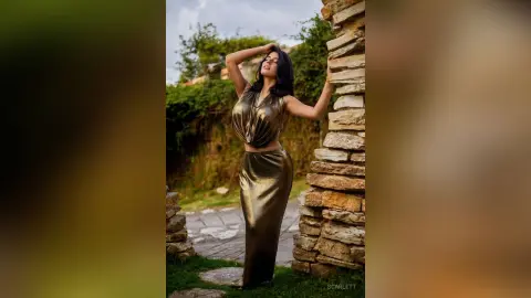 Elegant woman in a metallic gold satin gown posing by a rustic stone wall in a lush garden setting.