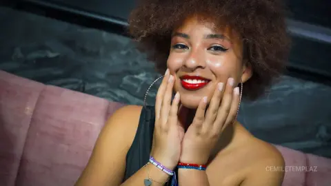 Smiling young woman with afro hair and red lipstick wearing a black top in a cozy lounge setting.