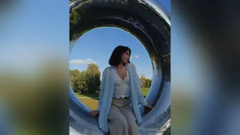 Young woman in a white top and light blue shirt relaxing outdoors near a large silver sculpture.