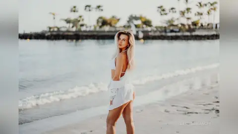 Blonde woman in a white summer outfit standing on a sunny beach with ocean waves in the background.
