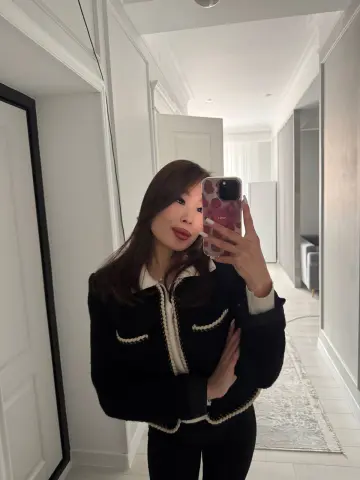Young woman in a chic black and white tweed jacket taking a mirror selfie in a bright modern hallway.