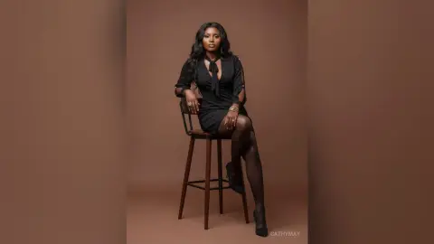 Elegant Black woman in a chic black dress sitting on a wooden stool against a minimalist brown studio background.