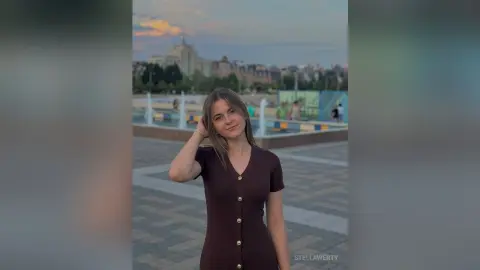 Smiling young woman in a brown button-down dress standing in a city plaza during a golden sunset.