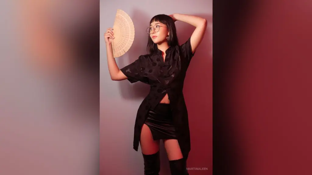 MartinaLeen β Stylish woman in a black button-down shirt and mini skirt holding a hand fan against a moody purple background.