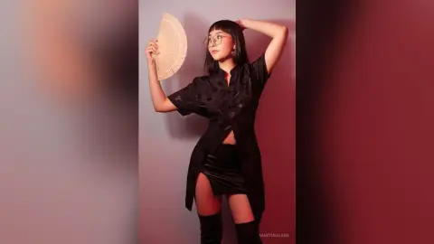 Stylish woman in a black button-down shirt and mini skirt holding a hand fan against a moody purple background.