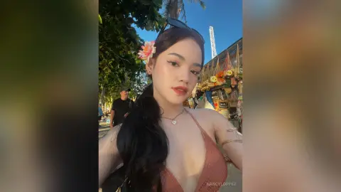 NancyLoppez poses outdoors wearing a peach bikini top with a pink flower in her long dark hair.