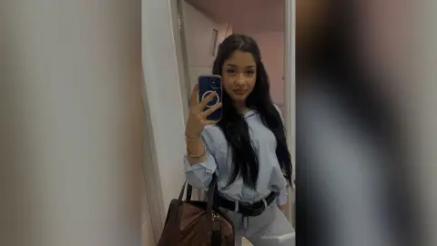 Young woman in a light blue button-down shirt taking a mirror selfie in a modern indoor setting.