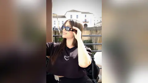 Smiling woman in a black t-shirt and sunglasses enjoying a sunny day on a balcony overlooking European architecture.
