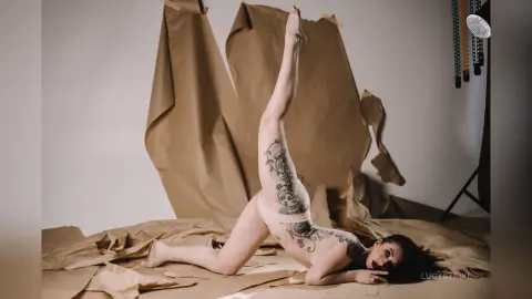 Tattooed woman in beige lingerie performing a yoga stretch against crumpled brown paper backdrop.