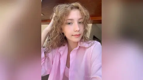 Young woman with curly blonde hair wearing a pink button-down shirt in a bright indoor setting.