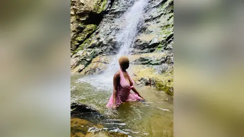 Woman in a pink dress standing in a natural waterfall pool surrounded by rocky cliffs in a serene forest setting.