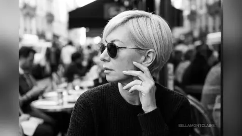 BellaMontclaire sits at an outdoor cafe wearing sunglasses and a dark sweater, looking off to the side.