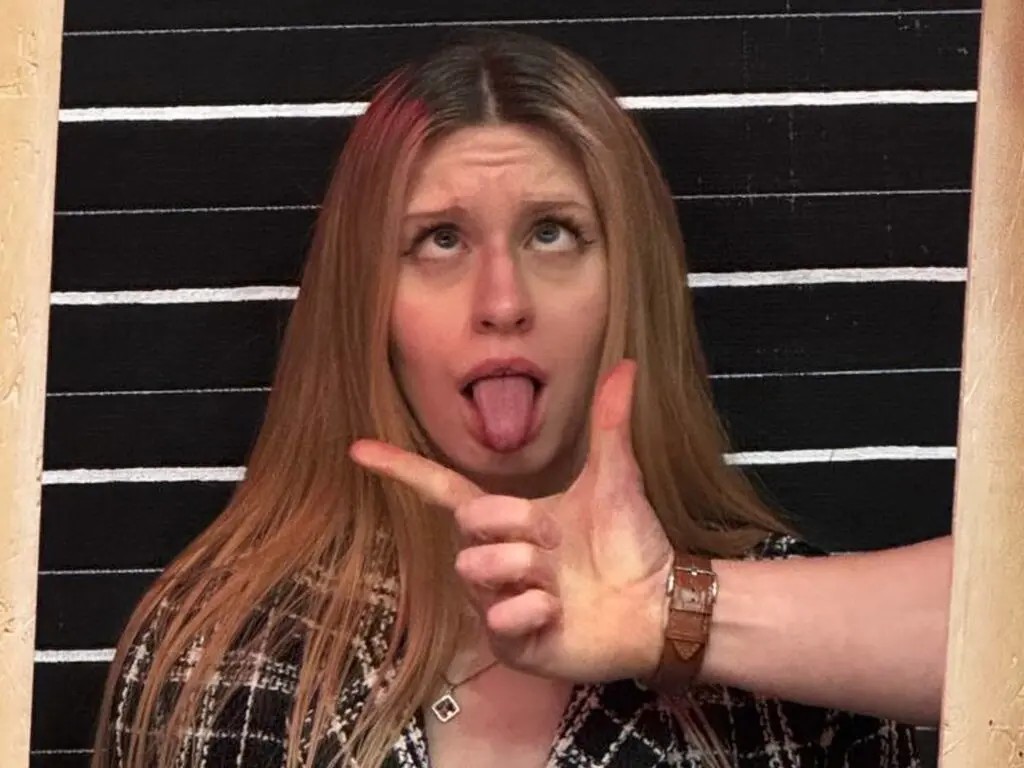 Playful blonde woman in a black plaid blazer making a funny face and hand gesture against a striped background.