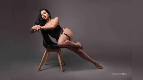 Glamorous woman in black lace lingerie posing on a chair against a moody grey studio background.