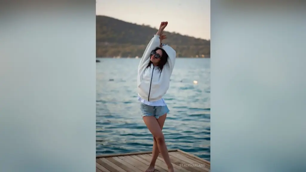 StacyHarland — Cheerful woman in a white sweatshirt and denim shorts stretching on a wooden pier by the ocean at sunset.