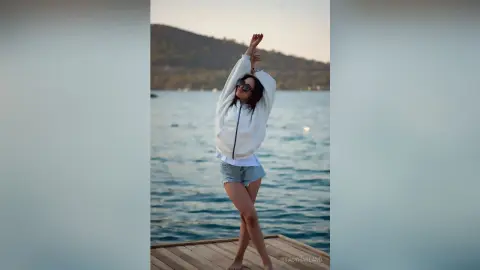 StacyHarland — Cheerful woman in a white sweatshirt and denim shorts stretching on a wooden pier by the ocean at sunset.