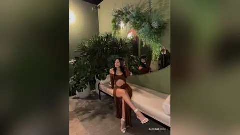 Woman in a brown cutout maxi dress sitting on a white sofa in a lush, green indoor garden setting.