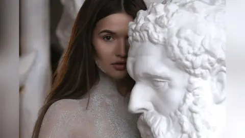 Young woman in a white lace dress posing next to a classical white marble bust in a bright, artistic setting.