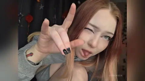Playful young woman with strawberry blonde hair and black nails making a peace sign in a casual grey sweatshirt.