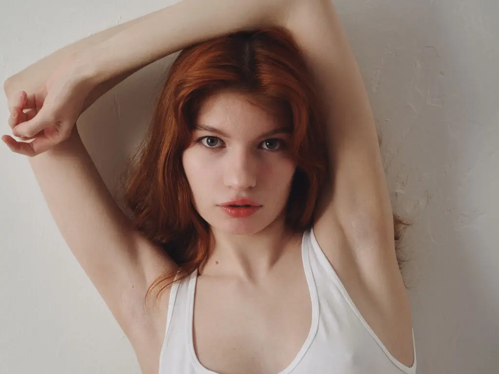 Redheaded woman in a white tank top posing against a white wall with a serene, minimalist mood.