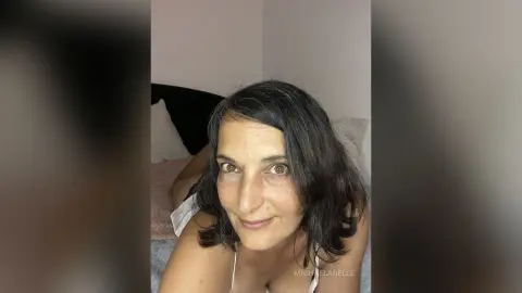 MichaelaBelle smiles while lying on a bed with her dark hair falling around her face.