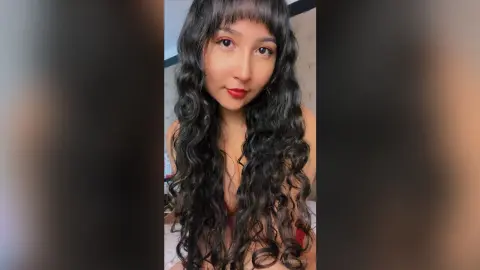 Young woman with long wavy dark hair and red lipstick posing in a cozy indoor setting.