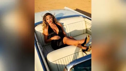 Glamorous woman in a black satin dress relaxing in a vintage white car seat under warm desert sunlight.
