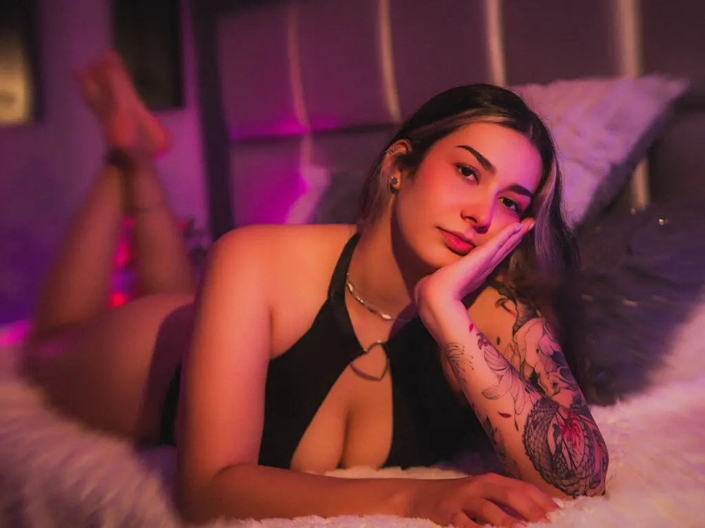 Young woman in a black bodysuit relaxing on a white faux fur bed in moody, warm pink lighting.