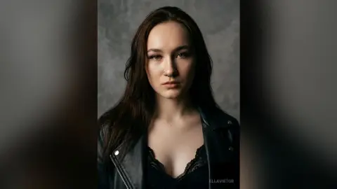 EllaVietor looks into the camera with long dark hair, wearing a black lace top and a black leather jacket against a grey background.